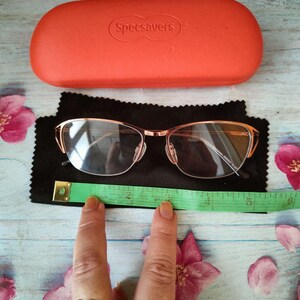 May include: Rose gold eyeglasses with an orange case from Specsavers. The glasses are on a black cloth with a green measuring tape. The case is rectangular and has the brand name printed on it. Pink flowers are scattered around the items.