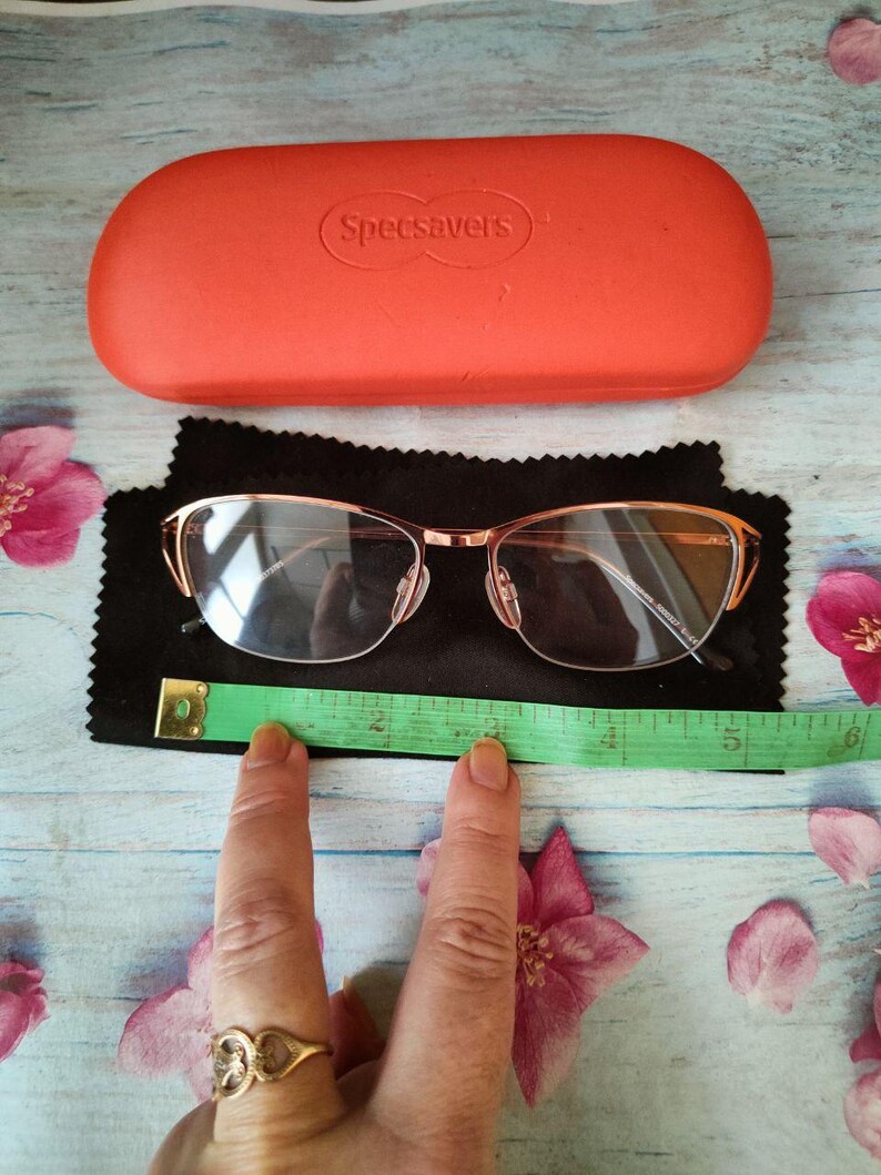 May include: Rose gold eyeglasses with an orange case from Specsavers. The glasses are on a black cloth with a green measuring tape. The case is rectangular and has the brand name printed on it. Pink flowers are scattered around the items.