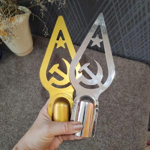 May include: Two metal flag pole finials, one in a gold finish and one in a silver chrome finish. Each finial features a cutout design of a hammer and sickle symbol topped with a five-pointed star within a pointed, teardrop-shaped frame.