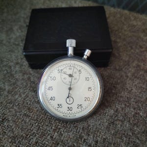 May include: A vintage stopwatch with a white face, black hands, and a silver metal casing. The stopwatch has a small dial and is marked with numbers from 0 to 60. The words "MADE IN USSR" are printed at the bottom. A black box is in the background.