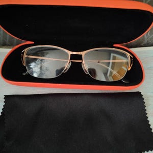 May include: Gold-framed spectacles in an orange and black case. A black cleaning cloth is below the case. The glasses have a modern, minimalist design. The case is open, displaying the glasses.