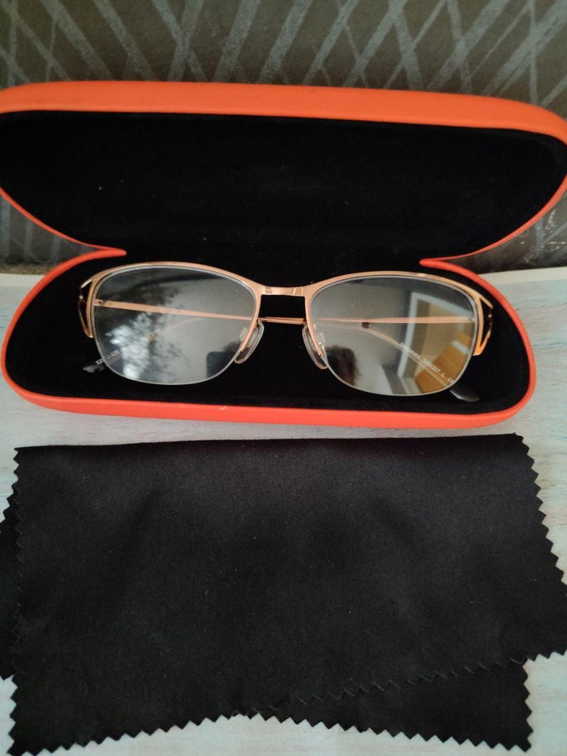 May include: Gold-framed spectacles in an orange and black case. A black cleaning cloth is below the case. The glasses have a modern, minimalist design. The case is open, displaying the glasses.
