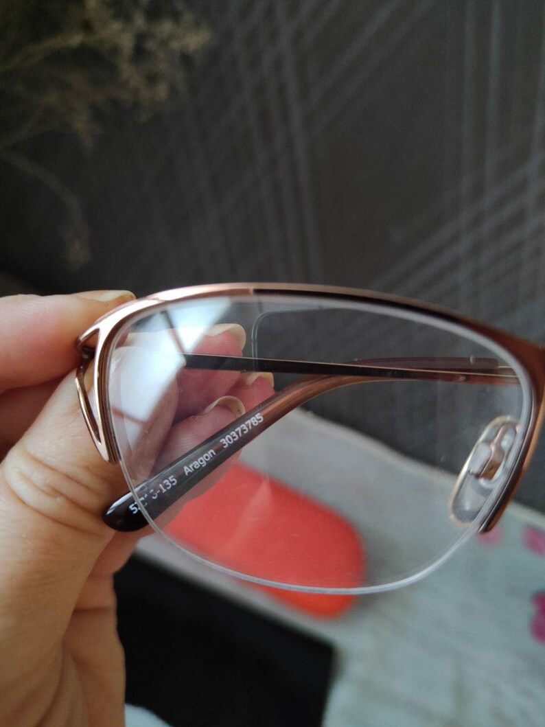 May include: Rose gold eyeglasses with a half-rim design. The glasses have clear lenses and a thin, delicate frame. The text "S-135 Aragon 30373785" is printed on the arm of the glasses.