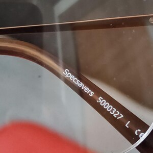 May include: Close-up of eyeglasses with brown frames. The text "Specsavers 5000327 L" is printed on the temple arm. The lenses are clear, and the frame has a subtle, metallic finish.