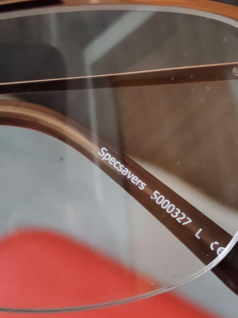 May include: Close-up of eyeglasses with brown frames. The text "Specsavers 5000327 L" is printed on the temple arm. The lenses are clear, and the frame has a subtle, metallic finish.