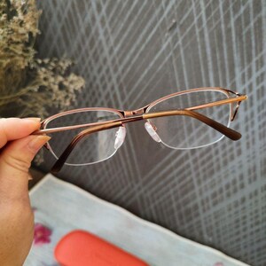 May include: Eyeglasses with a gold-coloured metal frame and clear lenses. The glasses are held by a hand, with a coral-coloured case and a black cleaning cloth in the background. The glasses have a half-rim design.