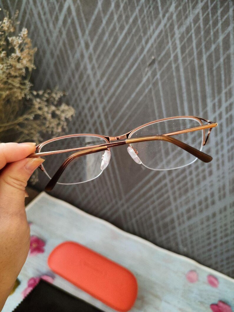 May include: Eyeglasses with a gold-coloured metal frame and clear lenses. The glasses are held by a hand, with a coral-coloured case and a black cleaning cloth in the background. The glasses have a half-rim design.