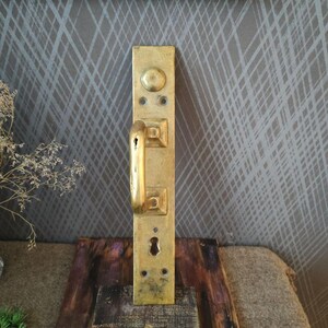 Peut inclure: Une poignée de porte en laiton vintage avec une plaque arrière rectangulaire. La poignée a un levier incurvé et un trou de serrure. Le laiton a un aspect vieilli et terni. La poignée de porte est montée sur une surface en bois.