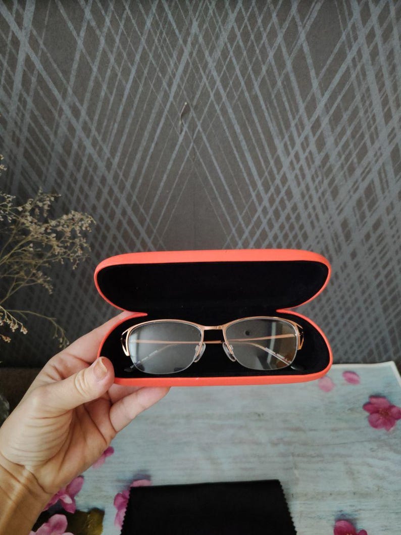 May include: Gold-framed spectacles with a rectangular shape are presented inside an open, bright orange glasses case. The case has a black interior. A black cleaning cloth is visible below the case.