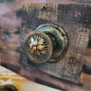 Könnte beinhalten: Nahaufnahme eines dekorativen, bronzefarbenen Schrankknaufs mit floralem Design. Der Knauf ist auf einer dunklen, strukturierten Holzoberfläche mit einem Used-Look-Finish montiert. Der Knauf hat eine zentrale Blume und einen detaillierten Außenring.