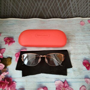 May include: A pair of gold-framed spectacles with clear lenses sits on a black cloth, beside a coral case with "Specsavers" printed on it. Pink flowers are scattered on a light blue wooden surface.