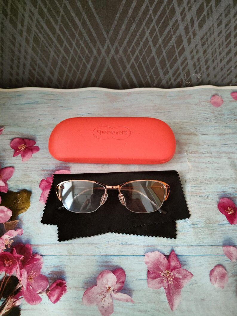 May include: A pair of gold-framed spectacles with clear lenses sits on a black cloth, beside a coral case with "Specsavers" printed on it. Pink flowers are scattered on a light blue wooden surface.