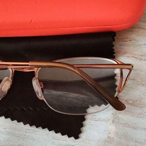 May include: Rose gold spectacles with clear lenses, placed on a black microfiber cloth. The glasses feature a thin, delicate frame and brown temple arms. An orange glasses case is in the background.