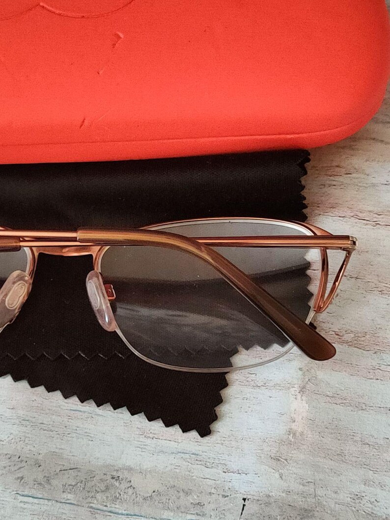 May include: Rose gold spectacles with clear lenses, placed on a black microfiber cloth. The glasses feature a thin, delicate frame and brown temple arms. An orange glasses case is in the background.