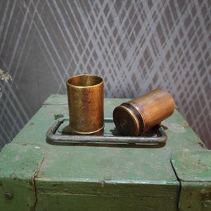 Peut inclure: Deux douilles cylindriques en laiton avec une finition métallique patinée. Une douille est posée verticalement, tandis que l'autre est couchée horizontalement sur le côté. Les deux objets présentent une surface lisse avec des traces d'oxydation.