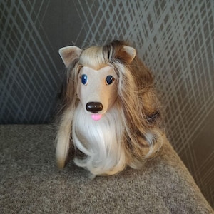May include: A plush collie dog toy with a tan and brown face, blue eyes, and long, flowing brown and white hair. The dog has a pink tongue and is sitting on a brown surface.
