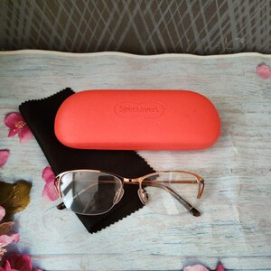 May include: Gold-rimmed spectacles with a coral case and a black cleaning cloth. The case is labelled "Specsavers". The glasses have a half-rim design. The case and glasses are on a light blue wooden surface.