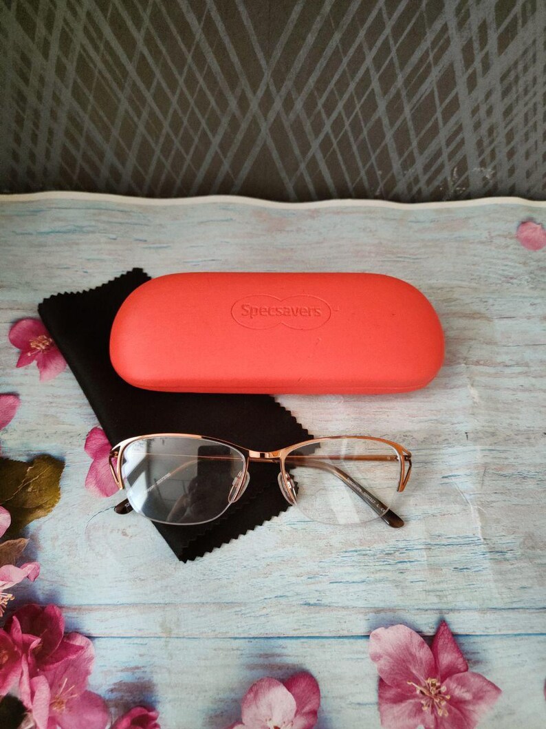 May include: Gold-rimmed spectacles with a coral case and a black cleaning cloth. The case is labelled "Specsavers". The glasses have a half-rim design. The case and glasses are on a light blue wooden surface.