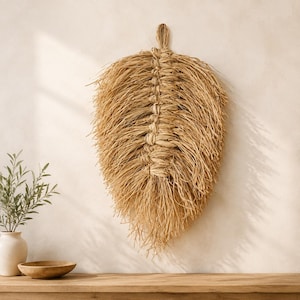 Könnte beinhalten: Ein Wandbehang in Form einer großen Feder, gefertigt aus natürlichen, beigefarbenen Bast- oder Strohfasern. Das Design besteht aus einem zentralen geflochtenen Mittelstrang mit langen, losen, ausgefransten Fasern, die eine strukturierte Form bilden.
