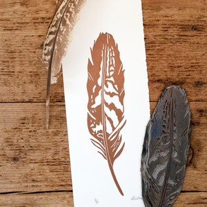 May include: A brown feather print on white paper with a woodblock carving of the same feather. The print is signed "Alethea".
