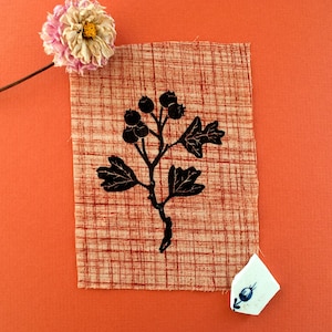 May include: A fabric square with a black botanical design, including berries and leaves, on a beige and red grid pattern. A dried flower and a piece of broken ceramic with a blue floral design are also present, set against an orange background.