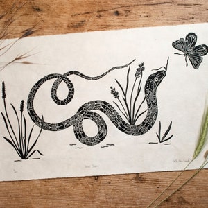 May include: Black and white linocut print of a snake with a butterfly above it. The snake is coiled around a group of tall grasses. The print is titled "Snake Charmer".