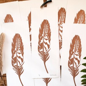 May include: Five brown prints of a single feather on white paper. The feather is detailed with a wood grain pattern. The prints are stacked on top of each other.