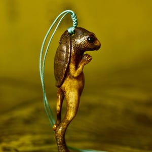 May include: A detailed, bronze-colored pendant in the shape of a turtle-like creature. The pendant has a textured shell and stands upright on two legs. A teal cord is attached for wearing. The text "波江 作品" is visible at the top.