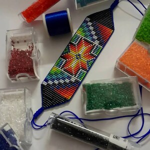 Puede incluir: Una pulsera de cuentas con un diseño geométrico colorido, con cuentas rojas, naranjas, amarillas, verdes y azules sobre un fondo negro. La imagen también muestra cuentas de varios colores en pequeños recipientes y una bobina de hilo azul.