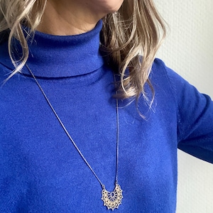 Könnte beinhalten: Eine goldfarbene Anhängerkette mit detailliertem, durchbrochenem Design. Die Kette liegt auf einem leuchtend blauen Rollkragenpullover. Der Anhänger hat eine halbkreisförmige Form mit filigranen Mustern.