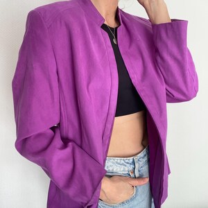 Könnte beinhalten: Eine leuchtend lila Jacke mit Stehkragen und durchgehendem Reißverschluss. Die Jacke ist aus einem glatten Stoff gefertigt und hat lange Ärmel. Die Jacke ist offen und zeigt ein schwarzes Oberteil und blaue Jeans.