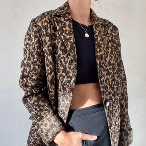 Könnte beinhalten: Ein brauner und goldener Leoparden-Blazer mit Knöpfen. Der Blazer ist aus einem strukturierten Stoff gefertigt und hat eine leicht verkürzte Passform.