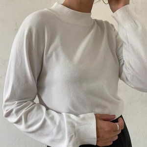 以下が含まれることがあります： リブ編みの襟とカフスが付いた、白い長袖のモックネックセーターです。
