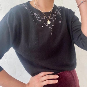 May include: A black sweater with a delicate floral design embroidered on the neckline. The sweater has a crew neck and long sleeves.