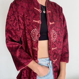 Può includere: Giacca bordeaux con motivo floreale e finiture rosa. La giacca ha un colletto alla coreana e chiusure a ranella decorative. Le maniche sono a tre quarti. La giacca è aperta, rivelando un top nero e jeans blu.