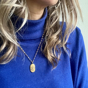 Könnte beinhalten: Eine goldfarbene Halskette mit einem detaillierten ovalen Anhänger hängt über einem leuchtend blauen Rollkragenpullover. Das Model hat blonde Haare.