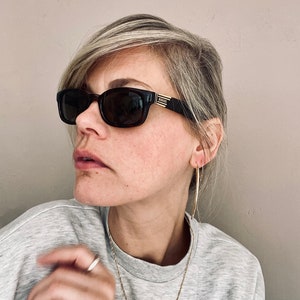 Könnte beinhalten: Eine Frau trägt eine schwarze Sonnenbrille mit goldenen Akzenten und dem Wort "VERSACE" auf dem Gestell. Sie trägt ein graues Sweatshirt und goldene Creolen.