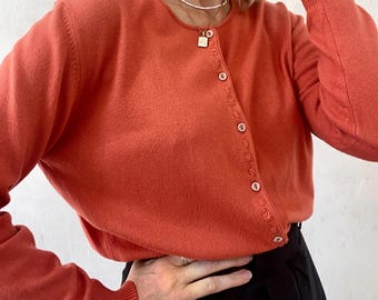 May include: A terracotta colored cardigan with a button-up front and long sleeves. The cardigan has a delicate stitch detail around the buttons.