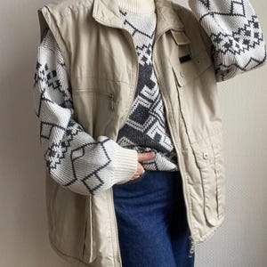 Op de afbeelding: Een beige vest met meerdere zakken en een ritssluiting. Het vest wordt gedragen over een trui met een geometrisch patroon in grijstinten en wit. De outfit bevat een blauwe jeans.