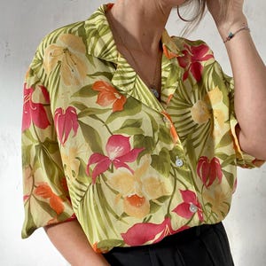 Puede incluir: Una camisa verde claro con botones y estampado floral tropical. La camisa presenta una variedad de flores en tonos de rosa, naranja y amarillo, así como hojas verdes.