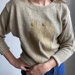 May include: A beige crew neck sweater with long sleeves. The front of the sweater has an embroidered design of two gold-coloured birds, with scattered rhinestone details. The sweater is made from a textured knit fabric.