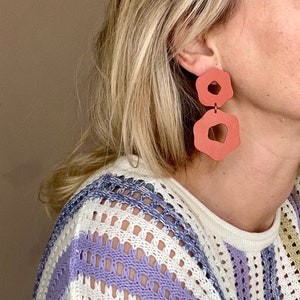 May include: A pair of terracotta-colored statement earrings with a unique, organic shape. The earrings are made of two pieces, one larger than the other, and are connected by a small metal loop.