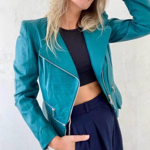 Könnte beinhalten: Eine tealfarbene Lederjacke im Biker-Stil mit Reißverschluss und silbernen Details. Die Jacke hat eine verkürzte Passform und eine leicht überdimensionierte Silhouette.
