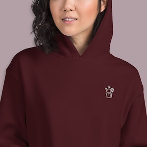 Pode incluir: Um moletom com capuz vinho com uma imagem bordada branca de uma cafeteira.