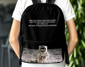 Może przedstawiać: Czarny plecak z białym nadrukiem astronauty na księżycu. Nadruk zawiera tekst "Here men from the planet Earth first set foot upon the moon July 1969, A.D. We came in peace for all mankind."