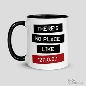 Könnte beinhalten: Weiße Keramiktasse mit schwarzem Rand und Henkel. Die Tasse hat schwarzen Text, der "There's no place like 127.0.0.1" in einem roten Kasten lautet.