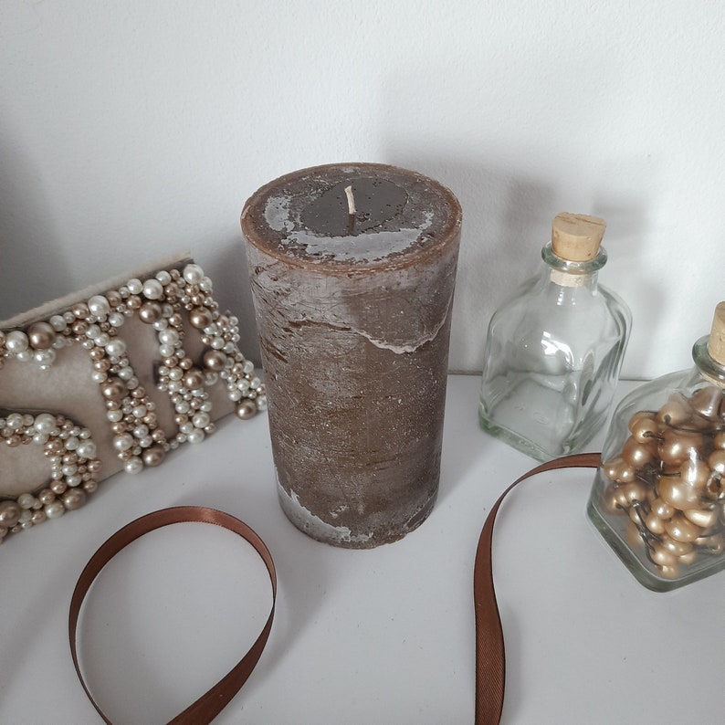 May include: A brown pillar candle with a textured surface. The candle is sitting on a white surface with a brown ribbon and a decorative beaded clutch.