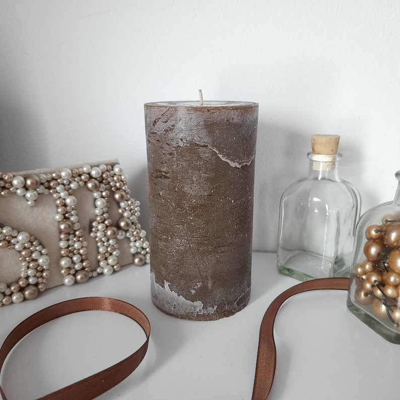 May include: A brown pillar candle with a white top and a rough, textured surface. The candle is sitting on a white surface.
