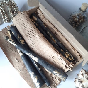 May include: A set of black taper candles with gold leaf accents, presented in a cardboard box with brown paper packaging. The candles are arranged at different angles, showcasing their textured surface and decorative design.