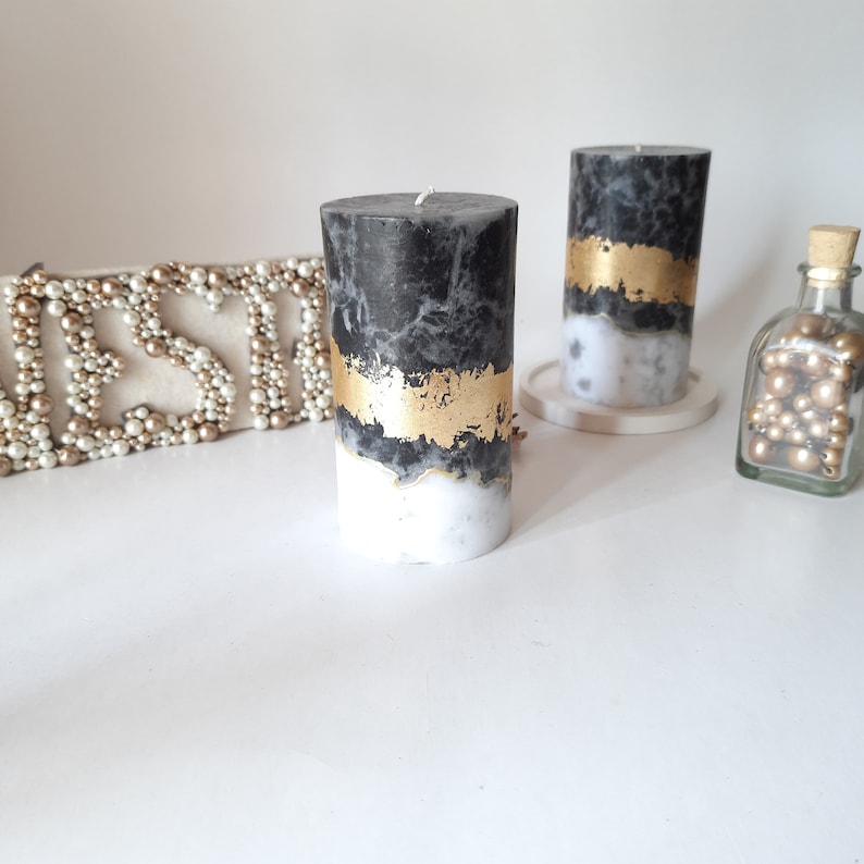 Puede incluir: Dos velas cil&iacute;ndricas con un patr&oacute;n de m&aacute;rmol en blanco y negro con una banda horizontal de pan de oro met&aacute;lico. Las velas tienen una sola mecha centrada en la parte superior y se muestran junto a una botella de vidrio llena de cuentas doradas.